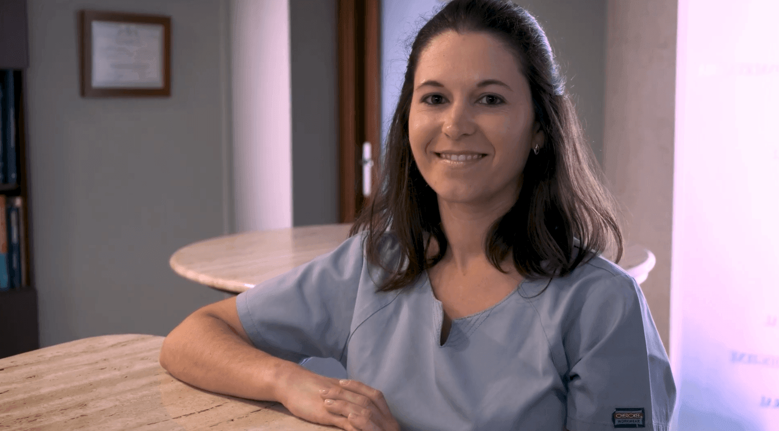 Smiling woman in scrubs leaning on a counter in a medical office setting.